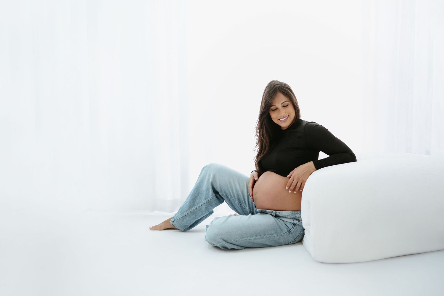 Artistic maternity photo with someone in casual denim against white wall.