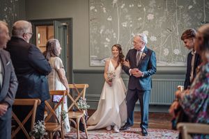 A wedding ceremony takes place in an elegant room with floral wallpaper and wooden chairs.
