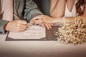 Close-up view of hands signing wedding documents with dried floral decorations and rings on a white surface.