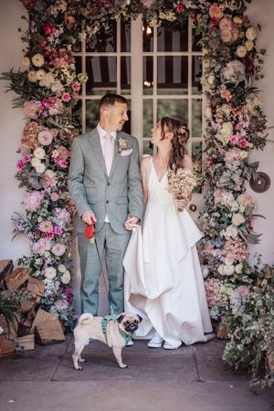 A couple poses with their pug under a stunning floral arch with pink and white roses at their wedding celebration.