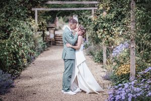 A couple walks through a garden path with purple flowers while holding their pug during a wedding photoshoot.
