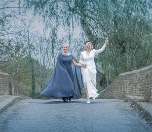 Two people in contrasting outfits walk along a stone wall path surrounded by willow trees.