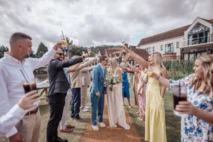 Wedding guests raise their glasses in a celebratory toast outside a waterfront venue on an overcast day.