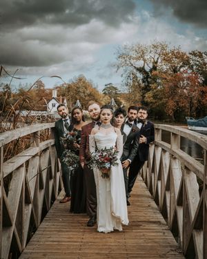 A wedding party poses on a wooden bridge under dramatic stormy clouds during an autumn day.