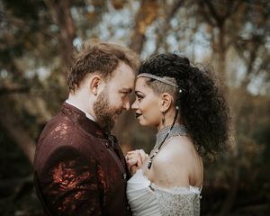 A romantic couple in wedding attire share an intimate moment outdoors with autumn trees in the background.