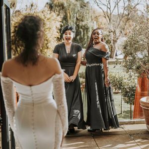 Wedding party members in dark dresses stand outdoors in golden sunlight while a bride in white looks on from behind.