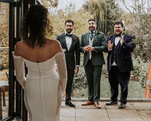 A wedding party stands together in formal attire outdoors with trees and natural lighting in the background.
