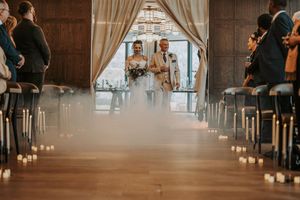 Wedding guests stand in an elegant rustic barn venue decorated with hanging lights and white curtains for a ceremony.