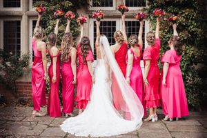 A group in bright pink bridesmaid dresses stand with a bride in white, raising their bouquets in celebration.