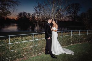 A romantic wedding couple poses at sunset near a lake with bare winter trees silhouetted against the sky.