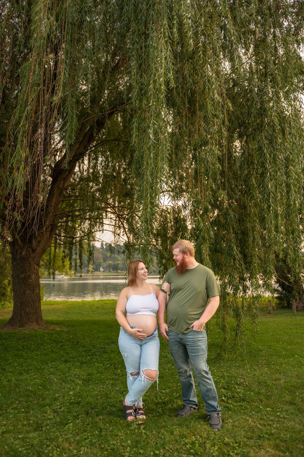 Expectant couple stands under weeping willow tree near lake for maternity photo.