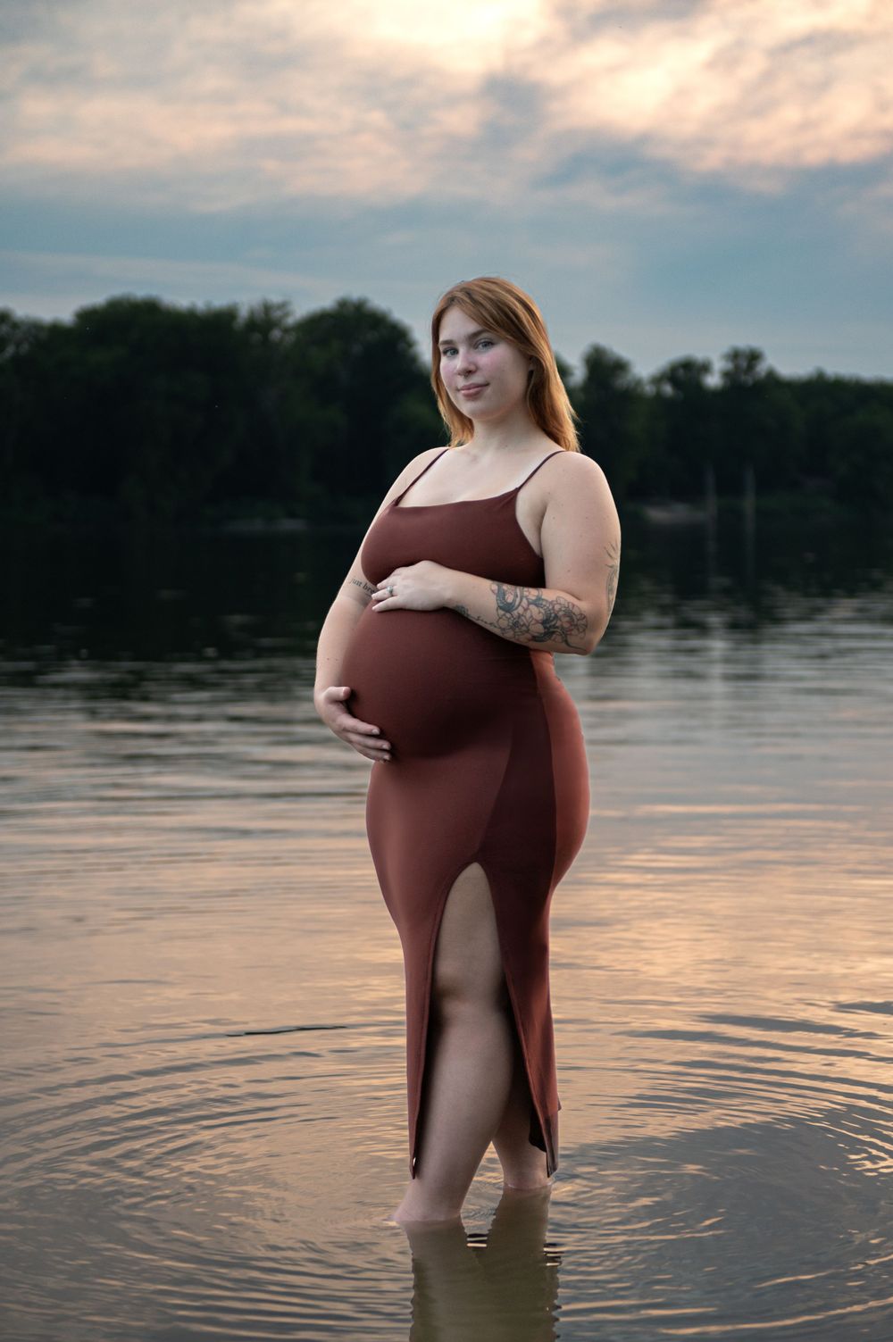 Expecting mother in burgundy dress wading in calm water during golden hour.