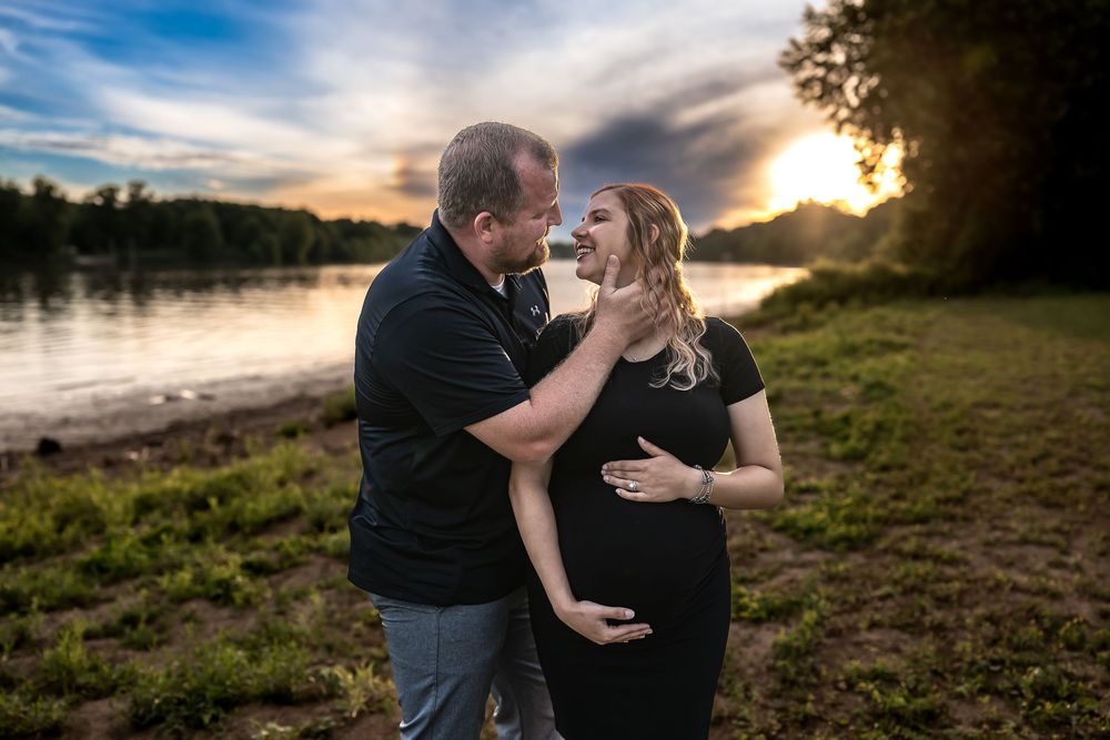 Couple sharing a tender moment during a maternity photo shoot by a river at sunset, woman in black dress.