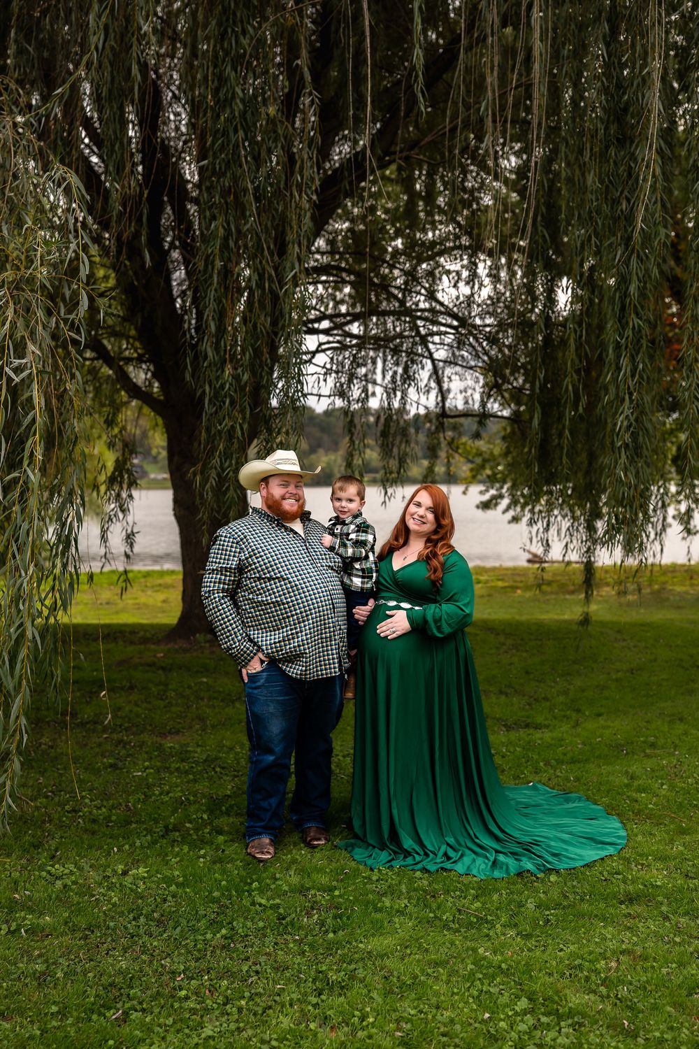 A family stands under a weeping willow tree on a grassy lawn, with the woman wearing an elegant emerald green gown.