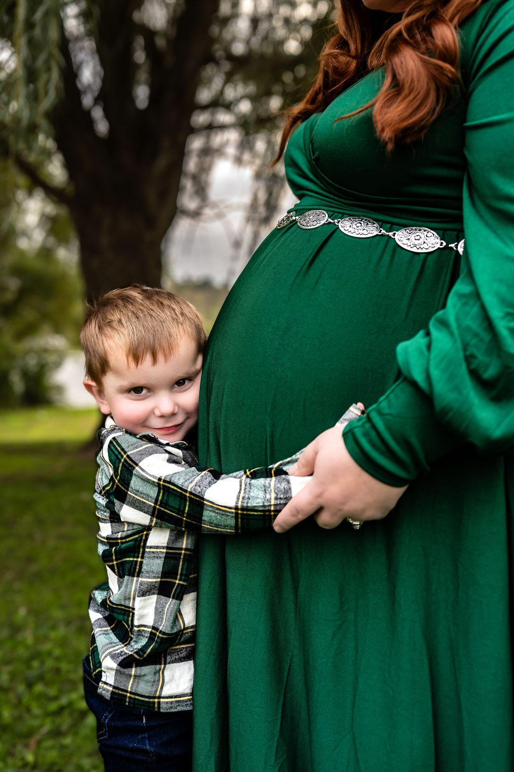 A young child in plaid shirt hugs a pregnant woman wearing an emerald green dress outdoors.