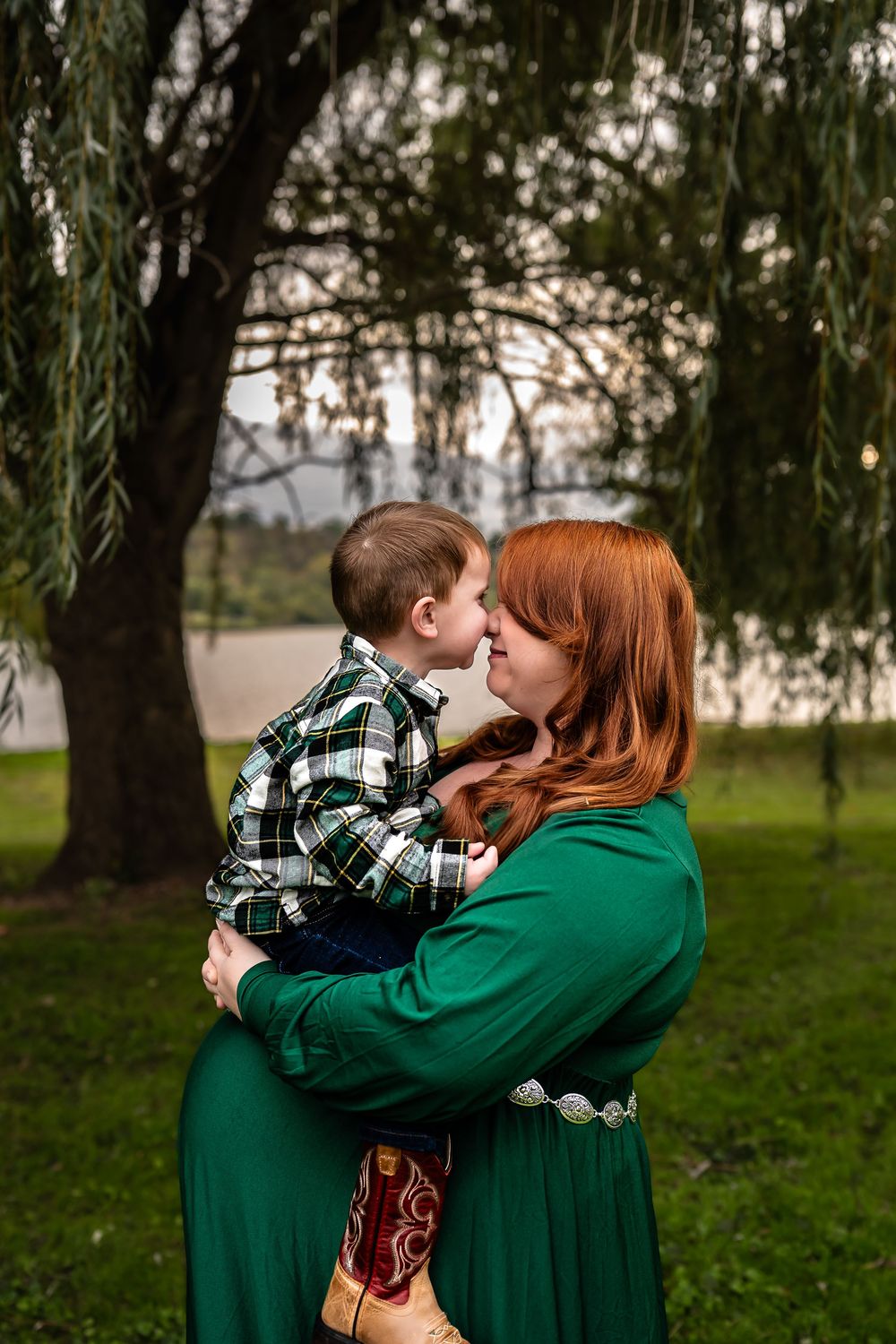 A couple wearing matching green outfits and plaid embrace tenderly under willow trees in a park setting.