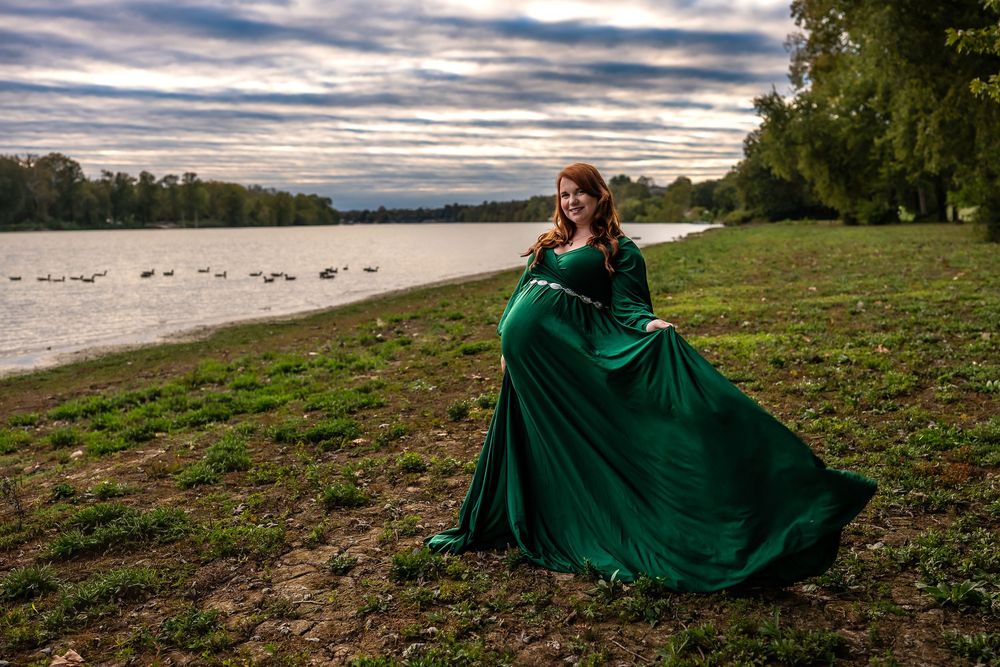 A flowing emerald green dress billows in the wind along a riverbank at sunset with dramatic cloudy skies.