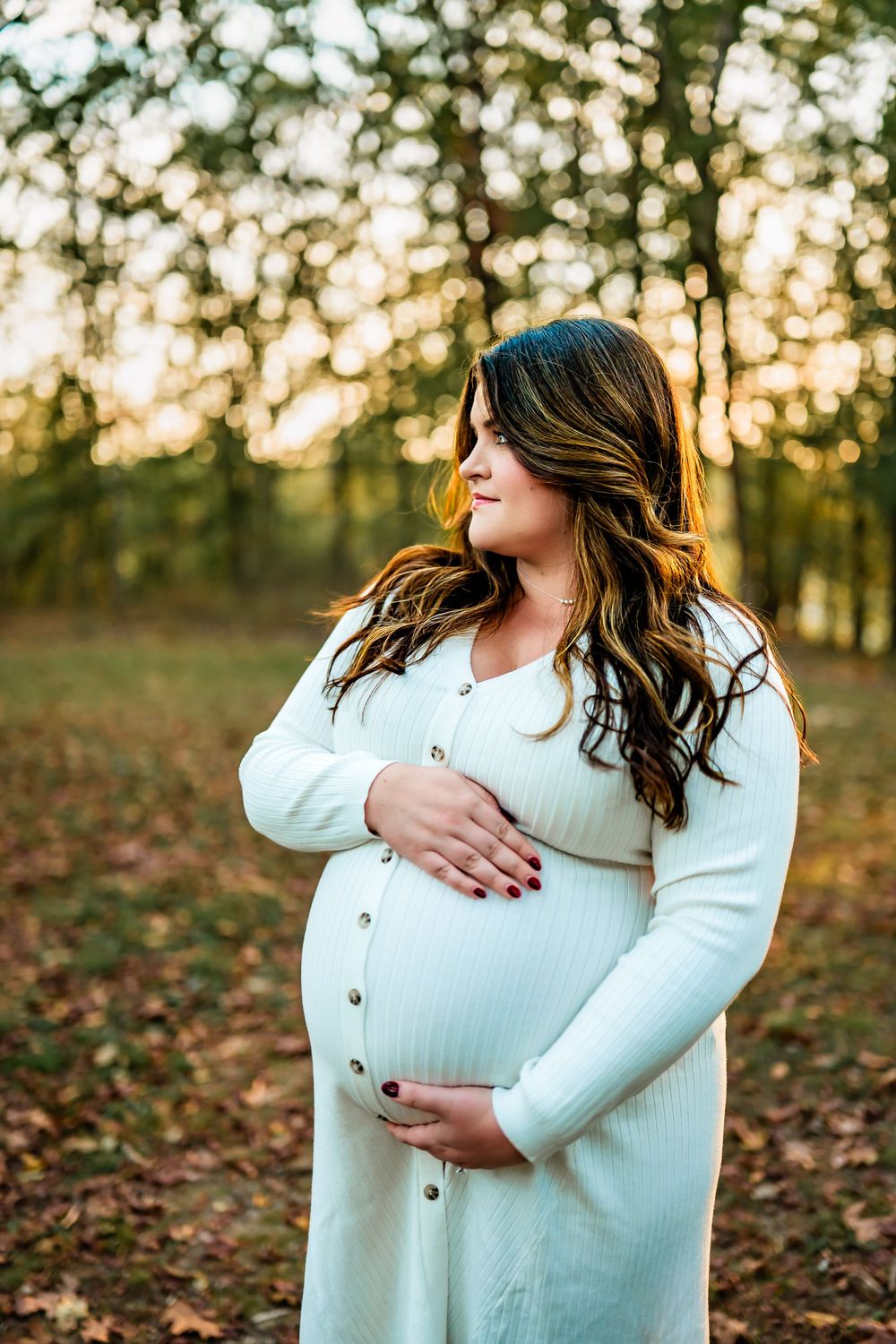 Pregnant woman in white sweater holds belly during autumn sunset maternity photoshoot in fallen leaves.
