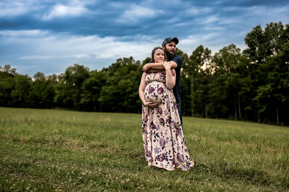 Pregnant couple poses in green field under dramatic cloudy sky, man embracing woman in floral maternity gown.