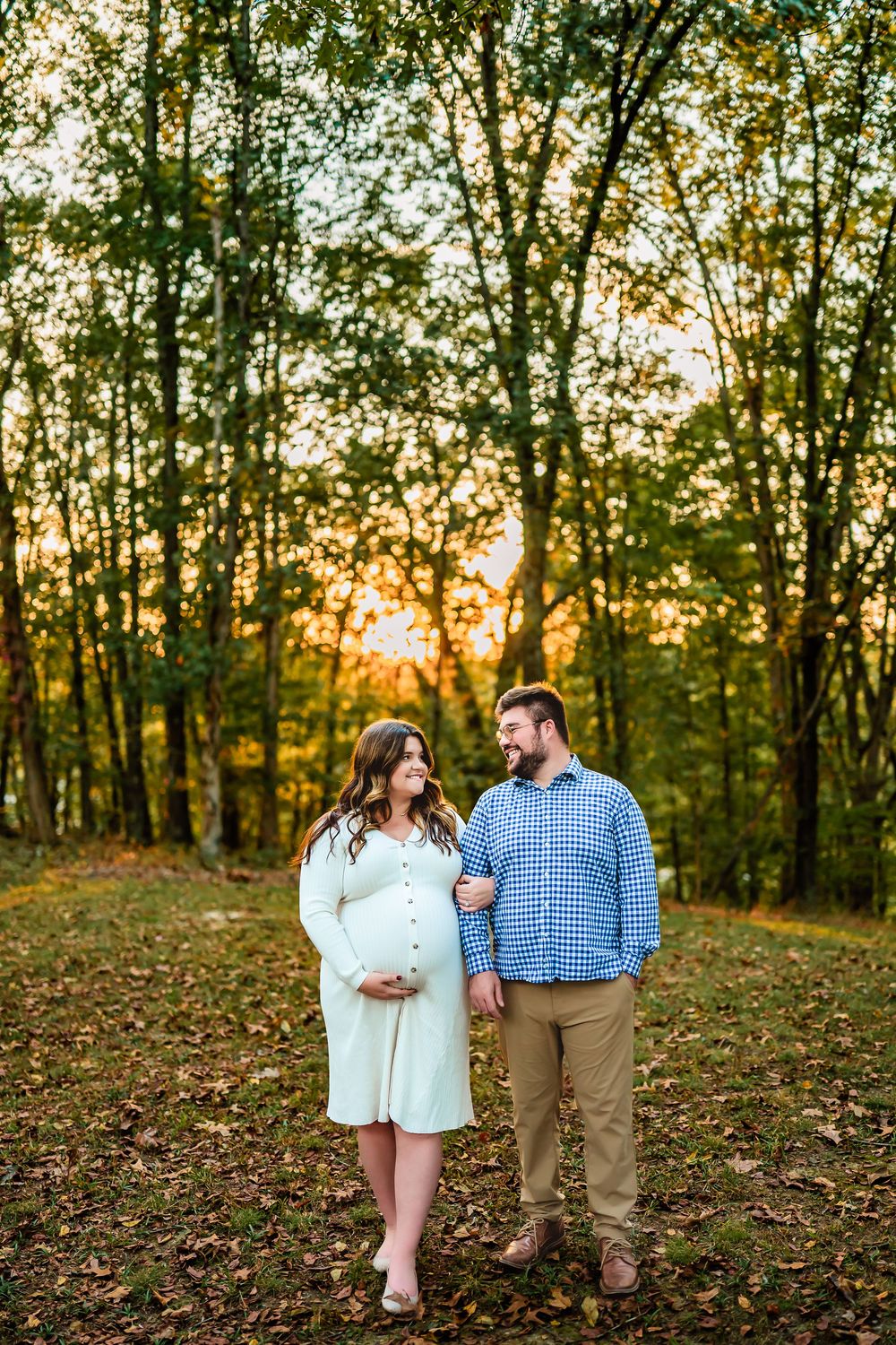 A couple stands together in an autumn forest at sunset with golden light filtering through the trees.