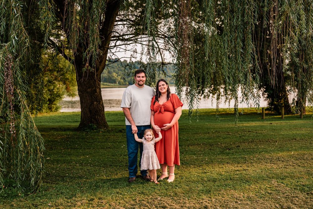 Pregnant woman in red dress poses with husband and toddler daughter under willow tree outdoors.
