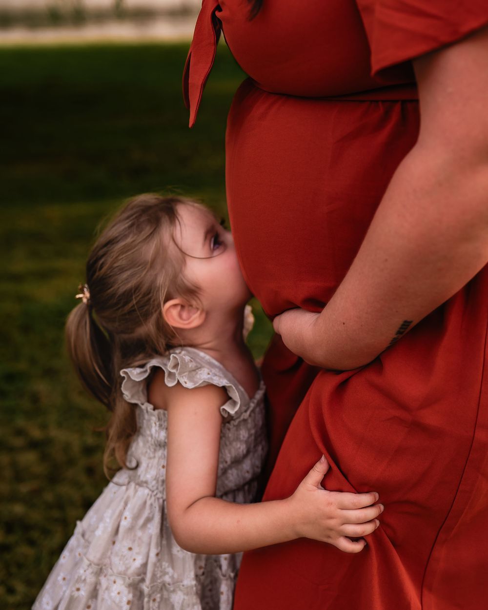 A young girl in a white dress hugs a pregnant woman in a red gown outdoors on green grass.