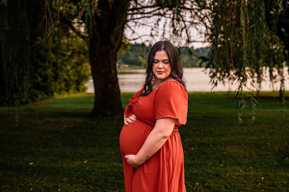 Pregnant woman in rust orange maxi dress cradling baby bump outdoors near willow tree and lake at golden hour.