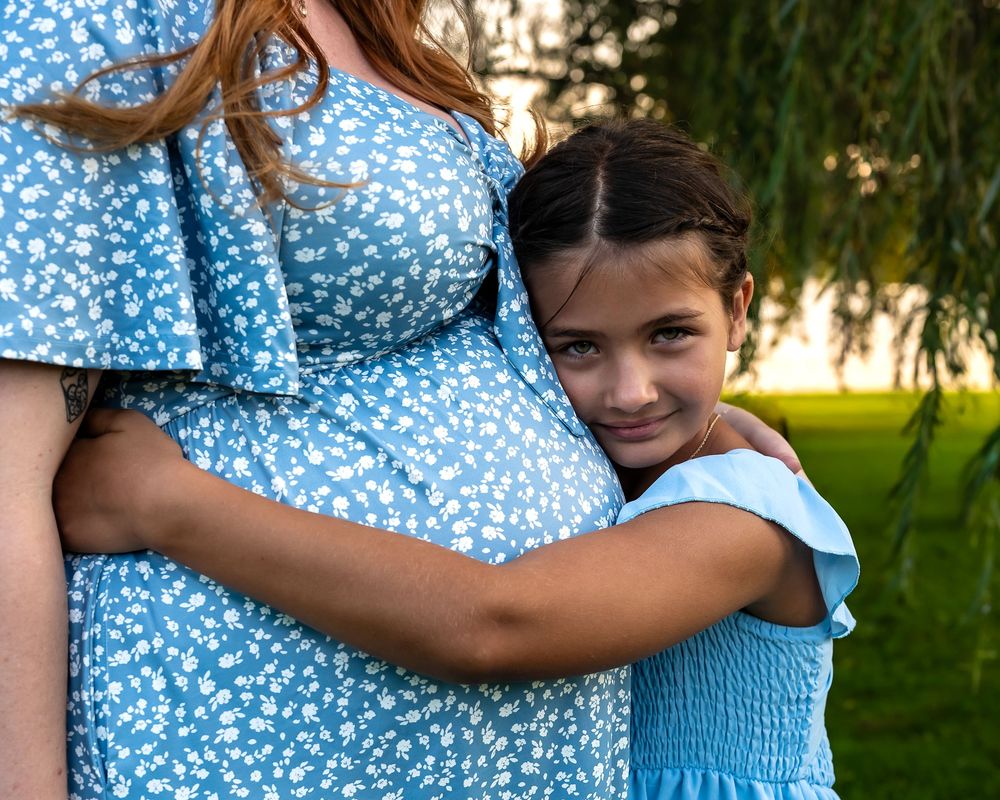 Young girl in blue dress hugging pregnant mother's belly outdoors near weeping willow tree.