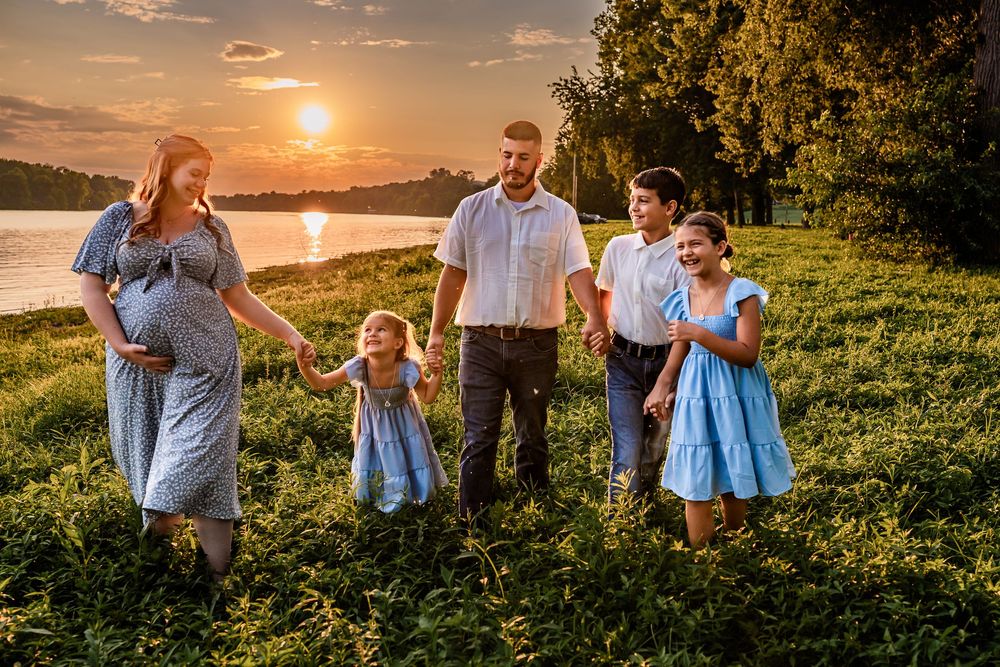 Pregnant woman and family holding hands at golden sunset by river, children in blue dresses, outdoor maternity photo.