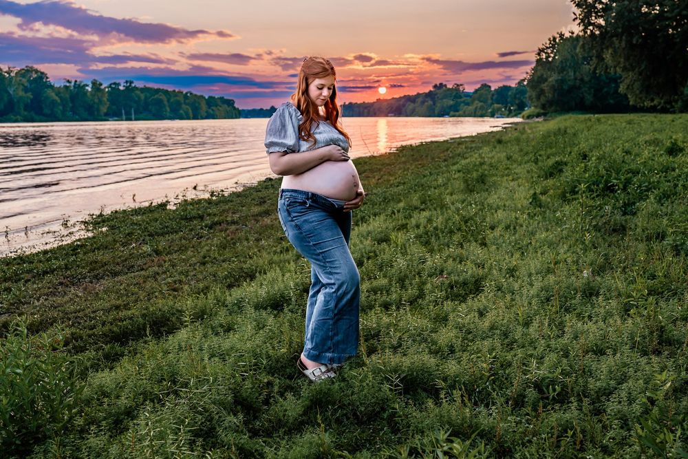 Pregnant woman cradling her baby bump by a serene river at sunset, wearing jeans and a blue shirt on green grass.
