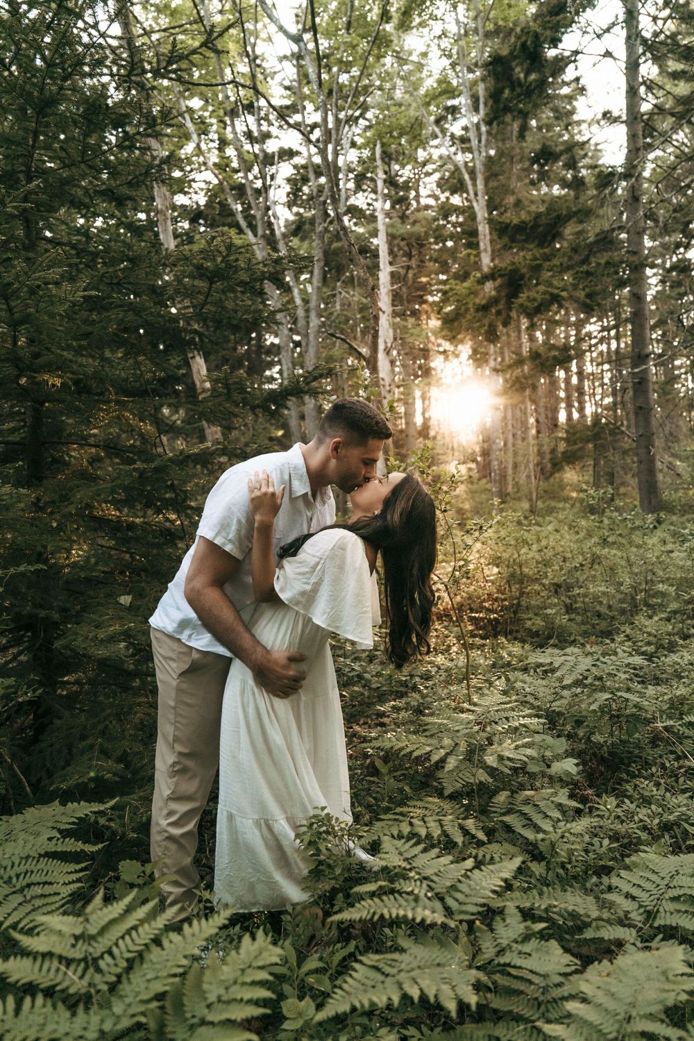 Romantic couple sharing intimate moment among ferns in forest with sunburst through trees.