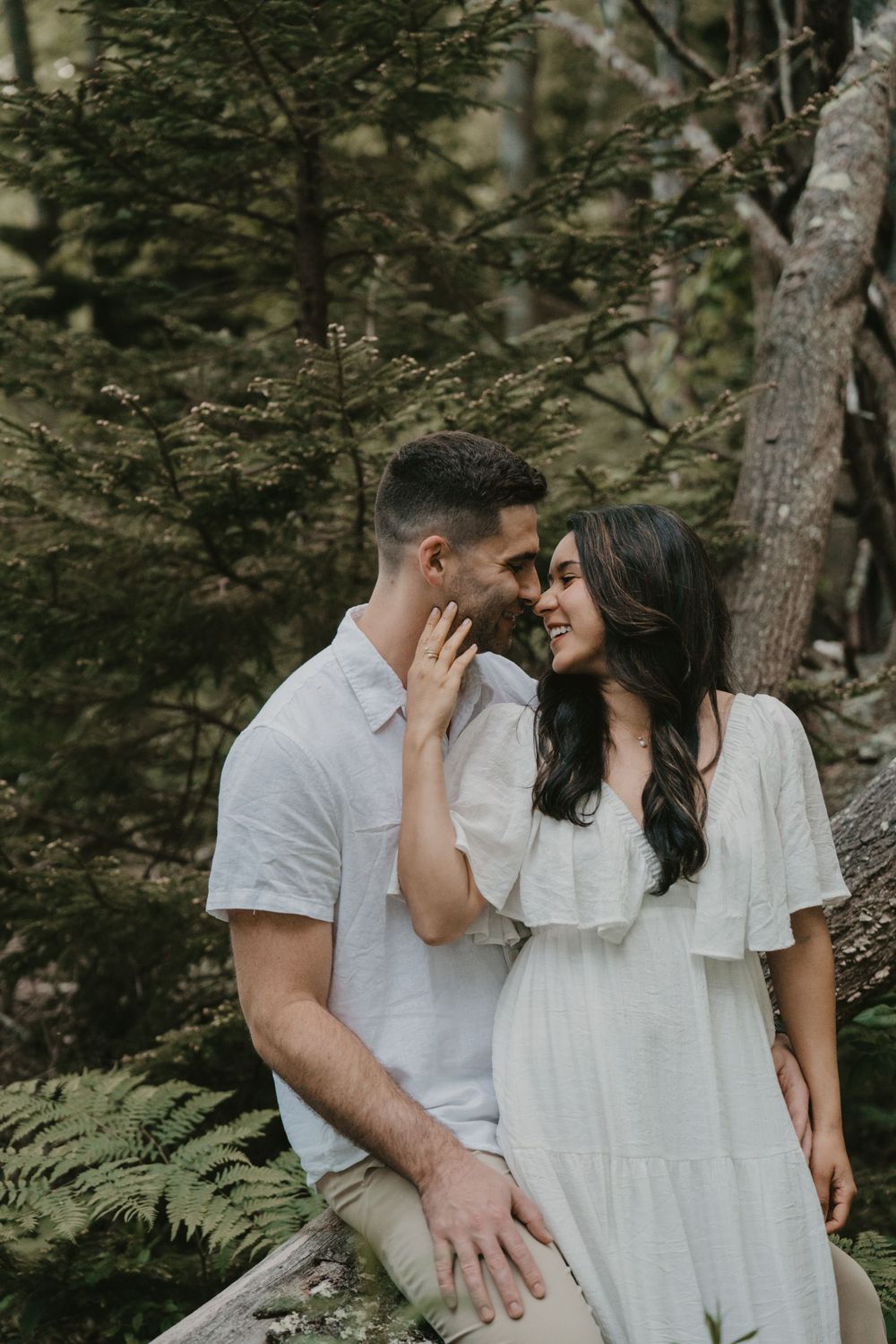 Couple in white outfits sharing tender moment against wooded forest background.