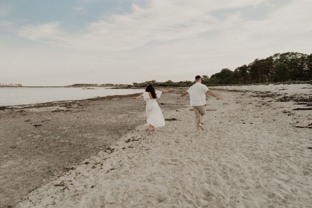 A couple walks hand in hand along a serene beach with trees in the distance.