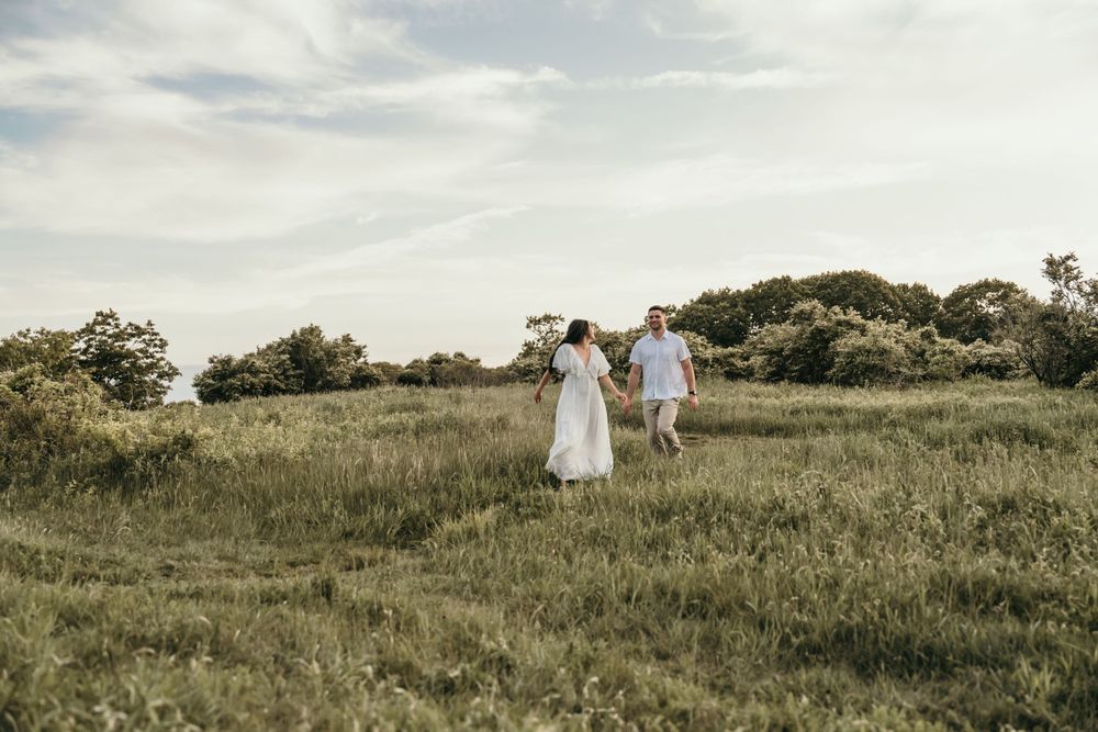 A couple walks through a grassy hilltop field during golden hour.