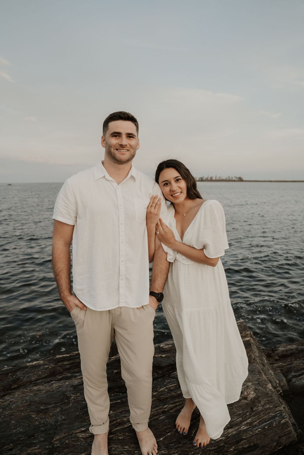 A couple poses together on rocky shoreline during coastal photoshoot.