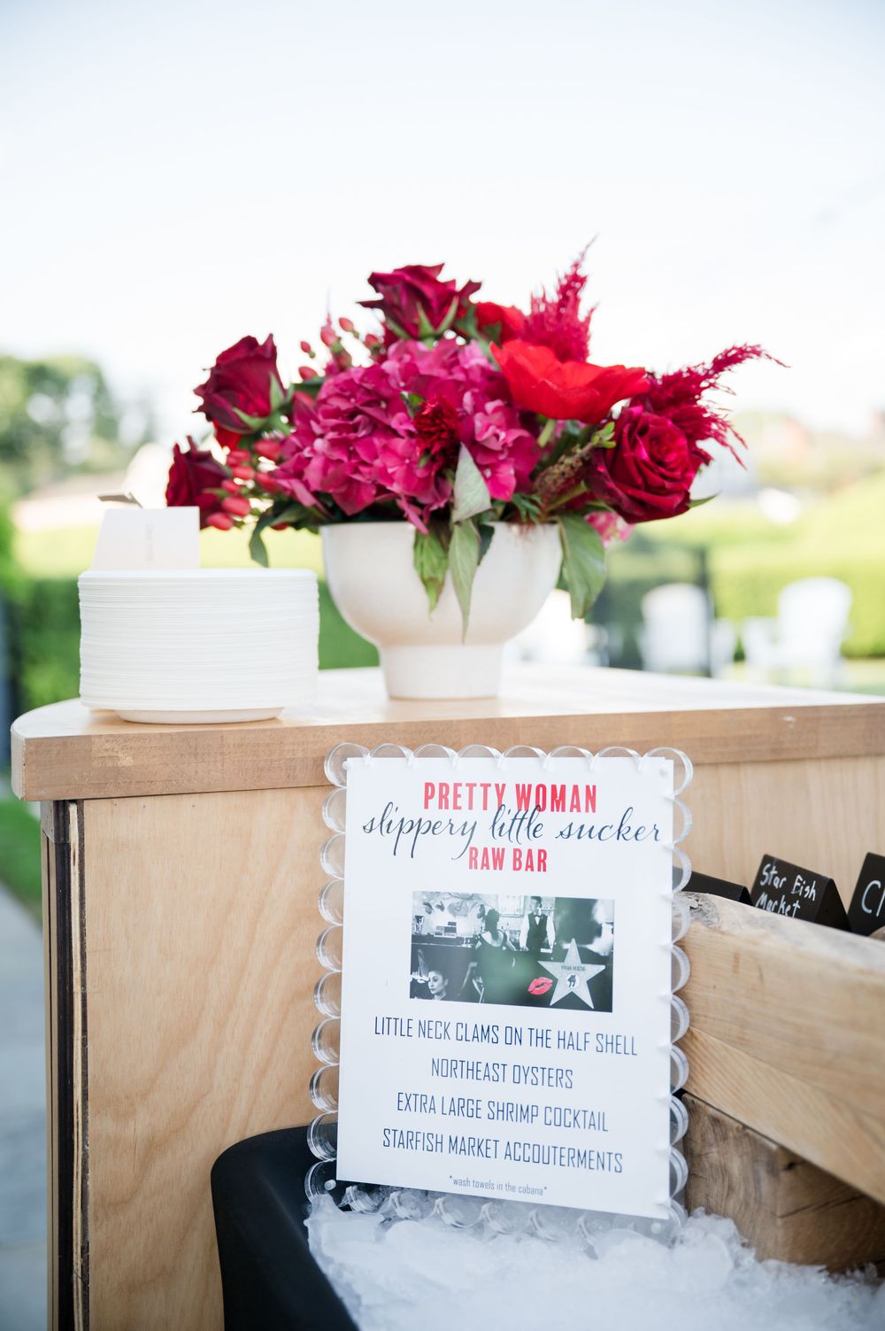 Wedding signage and red floral decorations displayed on wooden tables at an outdoor event.