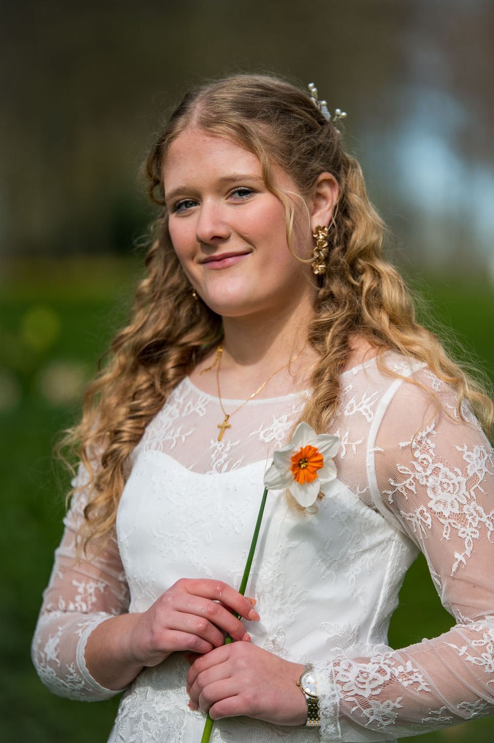 Young woman in white lace dress holding a daffodil flower outdoors in a garden setting.