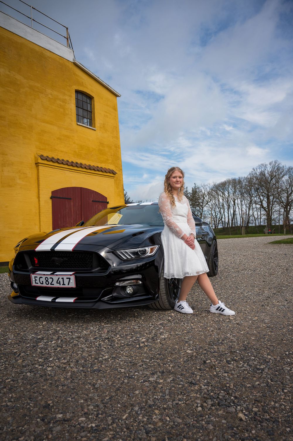 Young woman in white dress posing beside a black Ford Mustang with white stripes near yellow building.