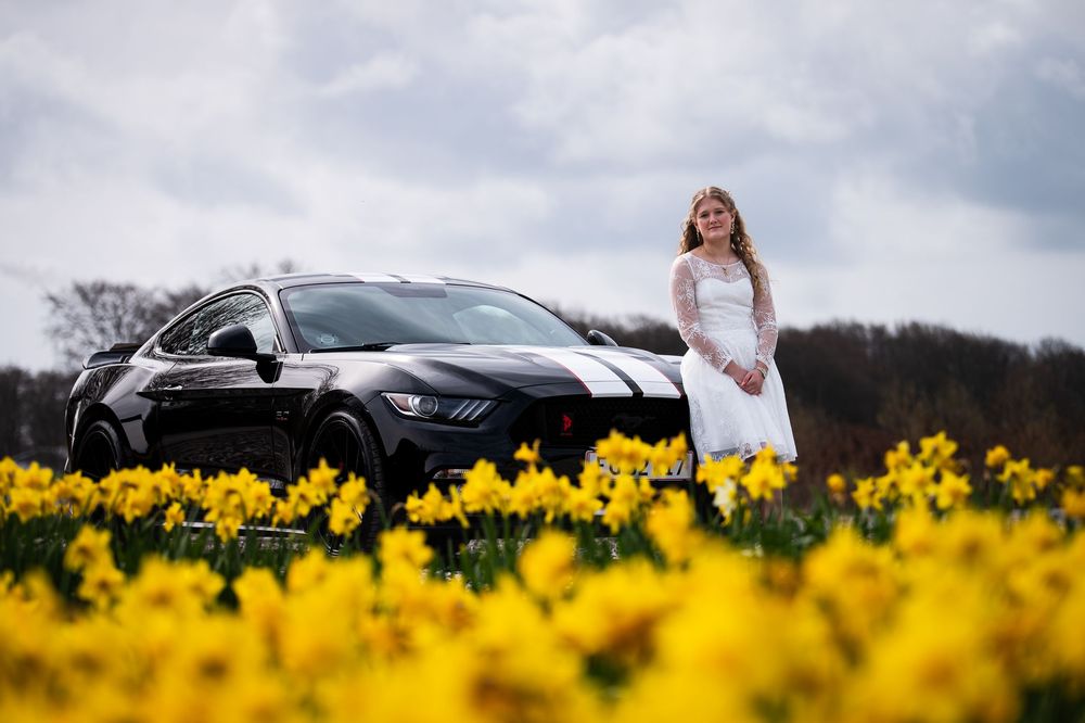 Young woman in white lace dress standing beside black Ford Mustang in a field of yellow daffodils.