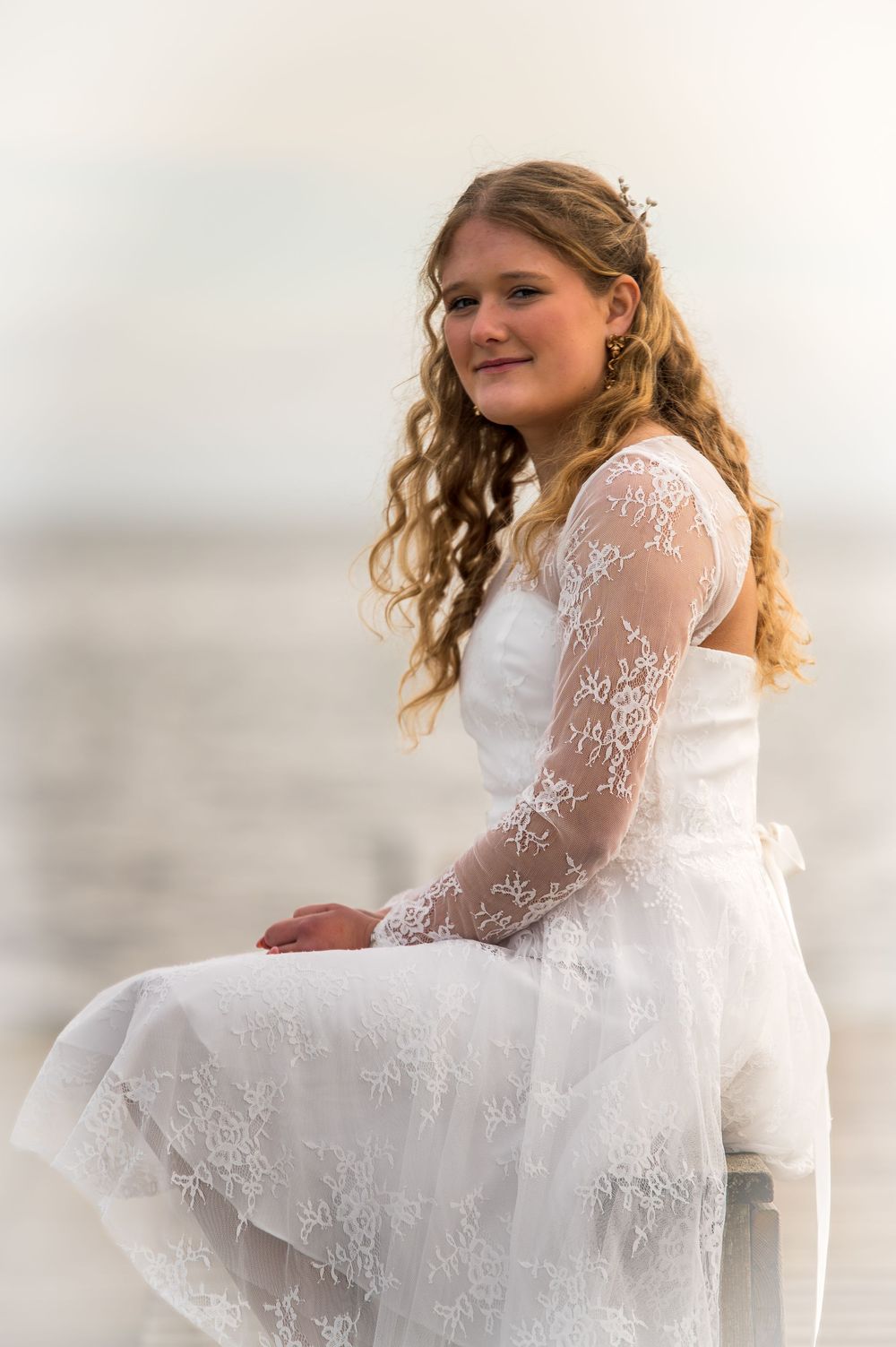 Young woman in elegant white lace dress sitting and smiling with soft water background behind her.
