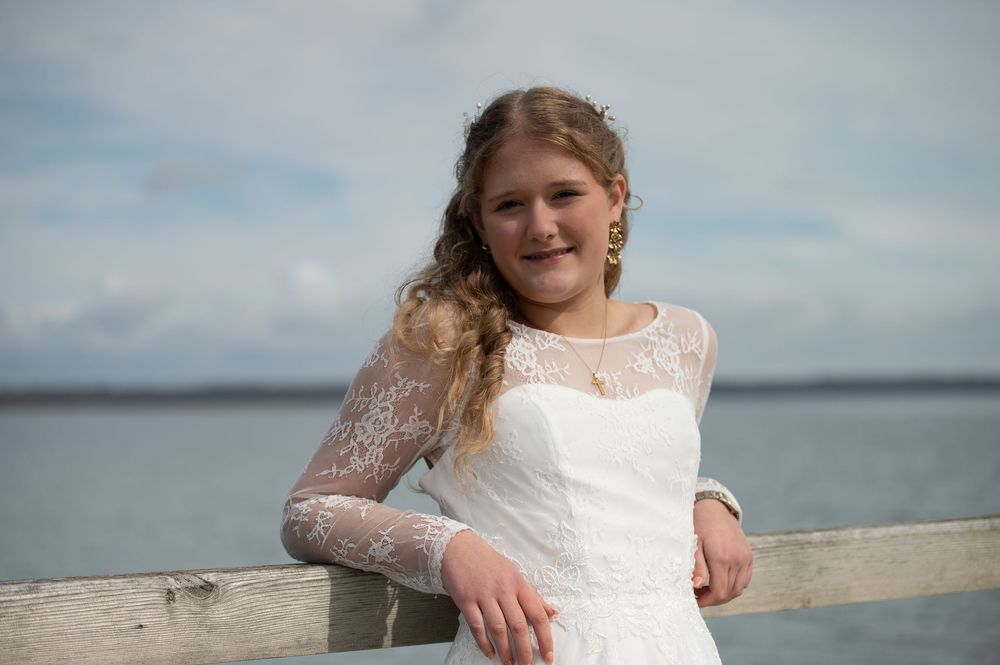 Young woman in white lace dress smiling while leaning on wooden railing with sea in background.