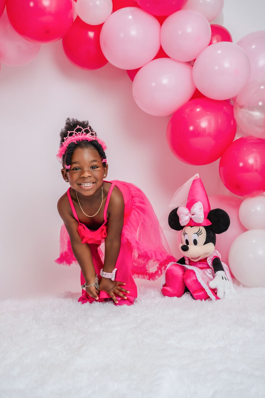 Young girl in pink tutu and tiara smiles beside Minnie Mouse plush, surrounded by pink balloon arch.