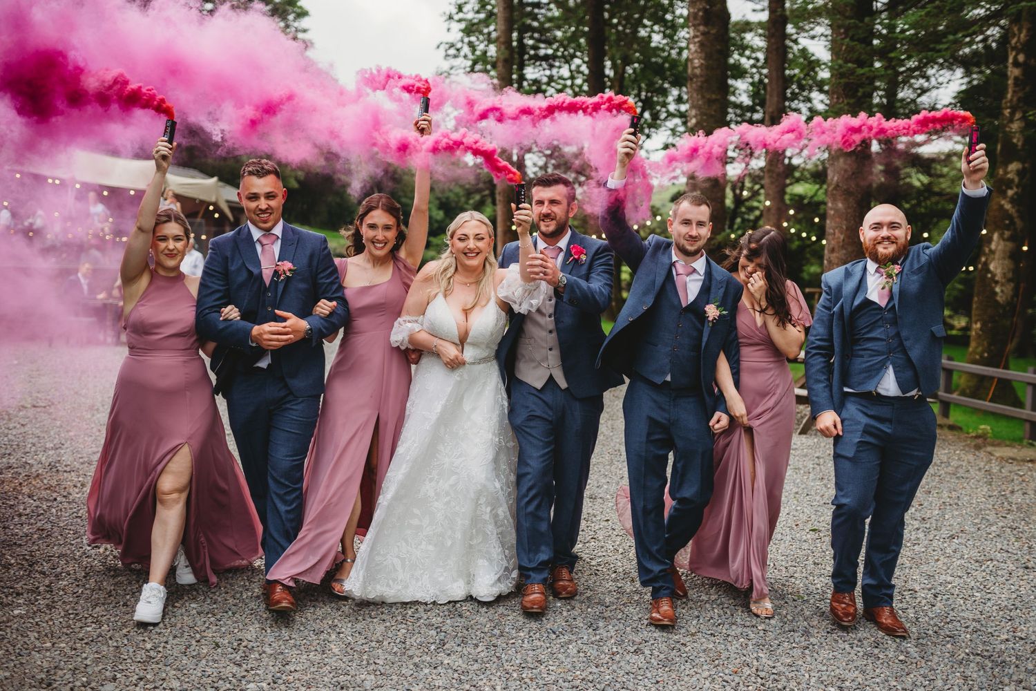 Wedding party celebrates with vibrant pink smoke bombs on a gravel path surrounded by trees.