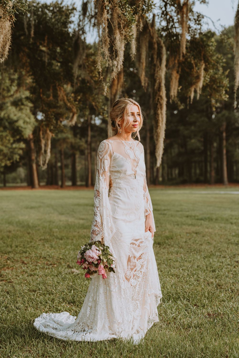 A lace wedding dress photographed in a sunlit field with Spanish moss draped trees.