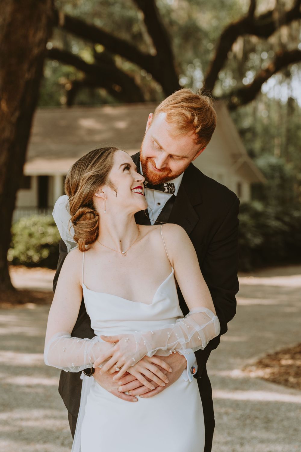 A tender wedding embrace photographed under oak trees in warm sunlight.