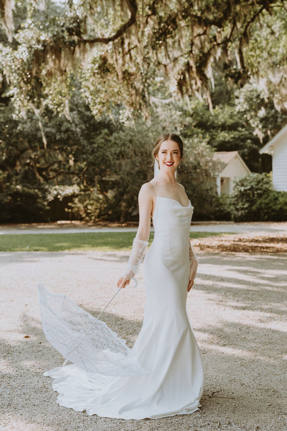 Bride in white gown twirls on outdoor path surrounded by moss-draped trees.