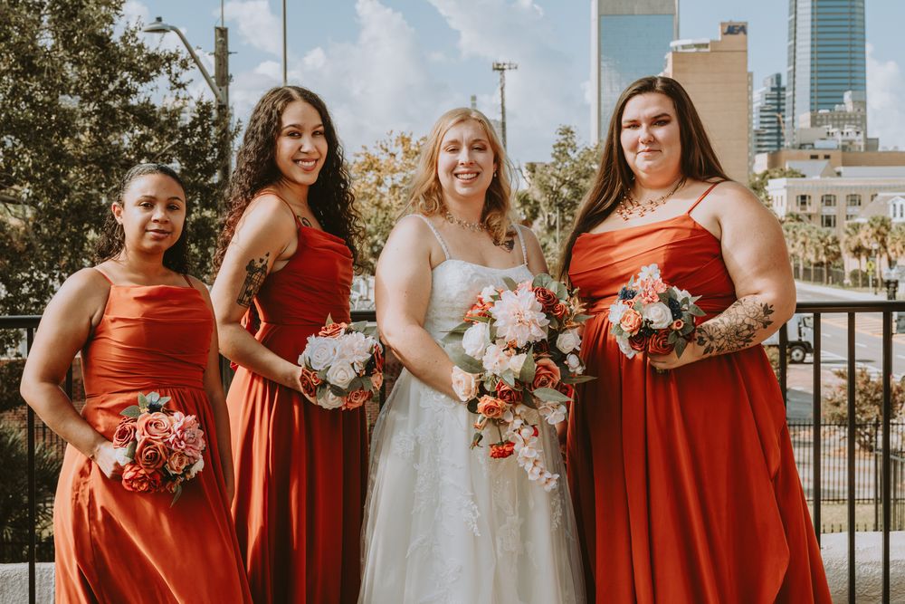 Bridal party wearing red dresses poses outdoors with bouquets against urban backdrop.