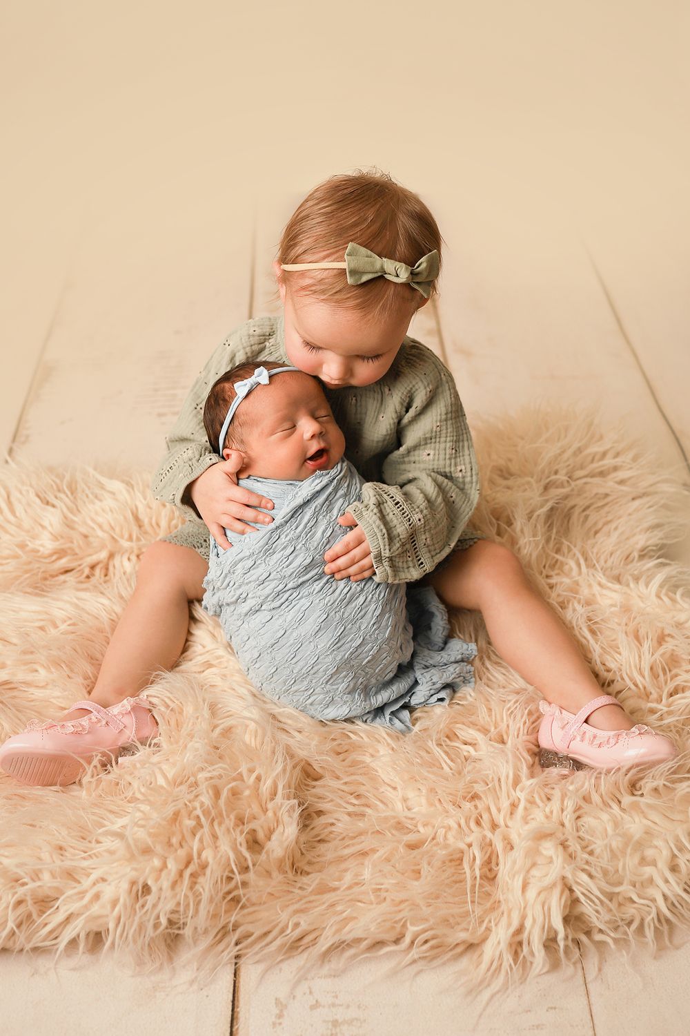 Toddler girl in green sweater hugging newborn sibling wrapped in blue on a fluffy beige rug.