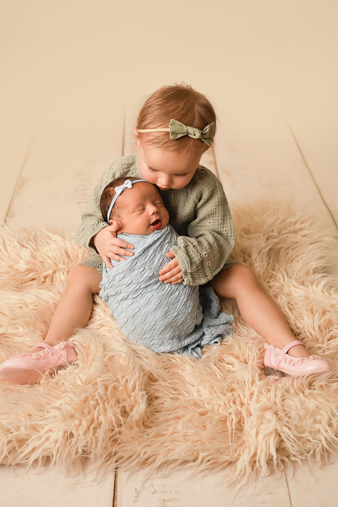 Toddler girl in green sweater hugging newborn sibling wrapped in blue on a fluffy beige rug.