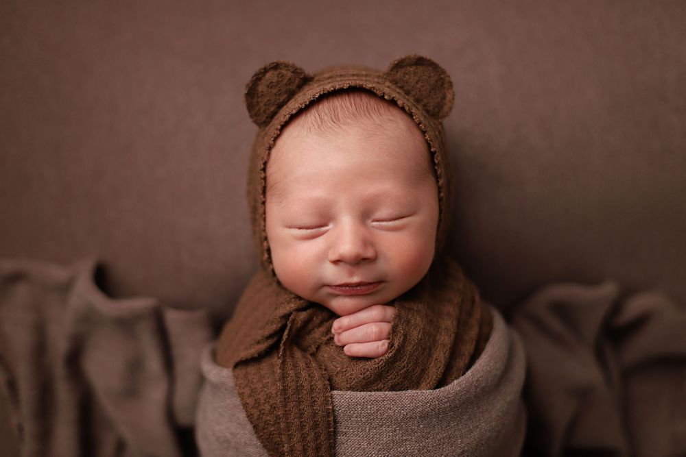 A sleeping baby wearing a brown bear-style bonnet and swaddled in cozy blankets against a dark backdrop. Utah newborn photography